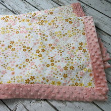 Load image into Gallery viewer, A folded baby blanket with a white background, covered in a delicate pink and yellow floral pattern, finished with a soft pink minky dot border, displayed on a rustic white-washed wooden surface.
