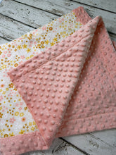 Load image into Gallery viewer, A folded baby blanket with a dusty pink minky dot texture on one side and a white floral print with yellow and pink flowers on the other, displayed on a rustic white-washed wooden surface.