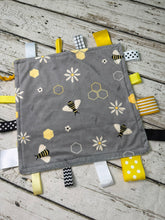 Load image into Gallery viewer, A square tag blanket baby sensory toy with a bumblebee and daisy pattern, featuring yellow, white, black, and gray ribbons on a rustic white wood background.