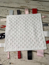 Load image into Gallery viewer, A square white minky dot baby lovey comforter with attached multi-colored fabric ribbon tags, displayed flat on a rustic white-washed wooden surface.