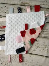 Load image into Gallery viewer, A baby sensory lovey blanket with a white minky dot fabric, a pink strawberry print and multiple colorful ribbon tabs on a rustic white wooden background.