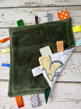 Load image into Gallery viewer, An olive green plush baby sensory blanket with dinosaurs surrounded by colorful patterned ribbon tags on a whitewashed wood background.