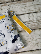Load image into Gallery viewer, A close-up of a fabric zip pouch with a white background and a gray and navy outdoor pattern, featuring a yellow dotted wrist strap with a silver metal clasp, set on a rustic white-washed wooden surface.
