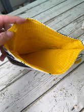 Load image into Gallery viewer, A hand holding open a small zippered pouch or bag made from yellow material, showing the bright yellow textured interior, set against a rustic white wooden table.