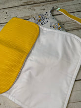 Load image into Gallery viewer, A reusable cloth diaper or changing pad shown on a rustic whitewashed wood surface. The product has a large white front panel, a bright yellow dotted flap, and a matching yellow strap with a metal clip.