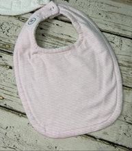 Load image into Gallery viewer, A light pink and white striped cotton bib with a soft curved shape, laid flat on a rustic whitewashed wooden surface.
