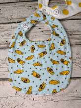Load image into Gallery viewer, A light blue fabric baby bib with an all-over print of Winnie the Pooh characters in yellow and brown, laid flat on a rustic white-painted wooden surface. A second bib with a yellow polka-dot pattern is partially visible at the top edge.