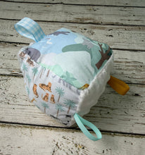Load image into Gallery viewer, A square fabric sensory toy or pillow with a woodland safari theme, featuring animal prints, palm trees, and a light blue gingham loop, resting on a rustic white-painted wooden surface.