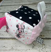 Load image into Gallery viewer, A handmade baby sensory toy cube featuring Minnie Mouse prints, pink and black polka dot panels, and a white hanging ribbon, resting on a distressed white wooden surface.