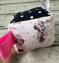 Load image into Gallery viewer, A handmade baby sensory toy cube featuring Minnie Mouse prints, pink and black polka dot panels, and a white hanging ribbon, resting on a distressed white wooden surface.