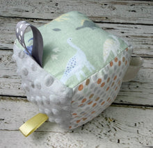 Load image into Gallery viewer, A handmade baby sensory cube toy with a pale green fabric top printed with dinosaurs, a gray minky dot side, a gray patterned ribbon loop