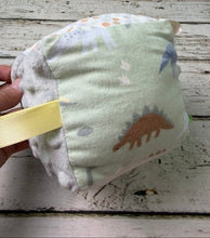 Load image into Gallery viewer, A handmade baby sensory cube toy with a pale green fabric top printed with dinosaurs, a gray minky dot side, a gray patterned ribbon loop