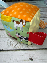 Load image into Gallery viewer, A small fabric baby activity cube toy with a farm theme, featuring an orange polka-dot top, a green side with cow, goat, duck, and pig illustrations, cream fabric sides, and a red ribbon tab, shown on a white wooden surface.