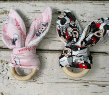 Load image into Gallery viewer, Two fabric teething rings with wooden rings, one pink with Minnie Mouse prints and one black, white, and red with Mickey Mouse prints, displayed on a rustic white wooden background.