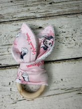 Load image into Gallery viewer, A pink Minnie Mouse themed baby teether rattle featuring plush bunny ears with a Minnie Mouse print, on a natural wooden ring handle, displayed on a rustic white-washed wooden background.