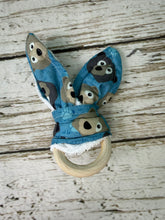 Load image into Gallery viewer, A fabric baby teething ring with bunny ears, featuring a blue pattern of bears and a natural wooden ring, displayed on a rustic white-washed wood background.