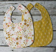 Load image into Gallery viewer, Two fabric baby bibs laid flat on a rustic whitewashed wood surface. The bib on the left is off-white with a pink, yellow, and beige floral pattern, and the bib on the right is gold yellow with a small white arrow motif.