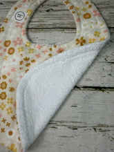 Load image into Gallery viewer, A close-up of a baby bib laid flat on a rustic whitewashed wood surface. The bib has a cream base with a small floral print in pink, yellow, and brown, and a soft white fleece or terry-like reverse side.