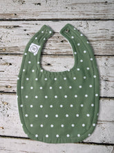 Load image into Gallery viewer, An olive green baby bib with a white polka dot pattern, laid flat on a rustic white-washed wooden plank background.