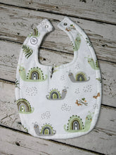 Load image into Gallery viewer, A white fabric baby bib with a playful print of green and gray snails under pastel rainbow arches, laid flat on a rustic, weathered gray wooden plank surface.