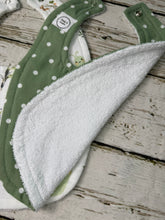 Load image into Gallery viewer, A close-up of a green baby bib with white polka dots and a soft white terry cloth lining, laid on a rustic whitewashed wooden surface.