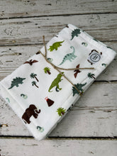 Load image into Gallery viewer, Two folded baby burp cloths displayed on a rustic white-washed wooden surface. The top one is white with a colorful dinosaur and palm tree print, and the bottom is sage green with white polka dots.