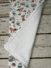 Load image into Gallery viewer, A folded baby burp cloth with a white plush terrycloth backing and a printed fabric cover featuring woodland animals like foxes, owls, and hedgehogs on a light background, displayed on a rustic whitewashed wood surface.
