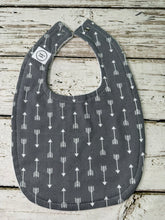 Load image into Gallery viewer, A dark gray fabric baby bib with a white arrow pattern, laid flat on a rustic white-washed wooden surface.