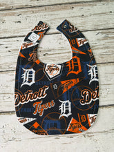 Load image into Gallery viewer, A baby bib featuring an all-over Detroit Tigers baseball print, laid flat on a light-colored rustic wooden background.