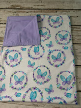 Load image into Gallery viewer, A folded baby blanket with a white background, decorated with a pattern of purple and blue butterflies inside floral wreaths, featuring a solid light purple reverse side, displayed on a rustic wooden surface.