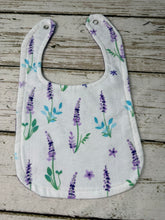 Load image into Gallery viewer, A white fabric baby bib with a printed pattern of purple lavender flowers and green leaves, laid flat on a rustic, weathered gray wooden surface.