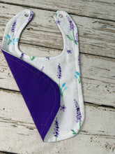 Load image into Gallery viewer, A fabric baby bib with a white front printed with purple lavender flowers and green stems, featuring a solid purple reverse side, laid flat on a rustic white-washed wooden surface.