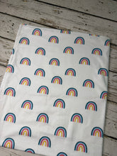 Load image into Gallery viewer, A white fabric blanket printed with a repeating pattern of colorful rainbows, laid flat on a rustic white-washed wooden surface.