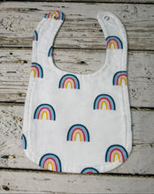Load image into Gallery viewer, A white fabric baby bib with a repeating rainbow pattern in blue, pink, and yellow, laid flat on a rustic, weathered gray wooden surface.