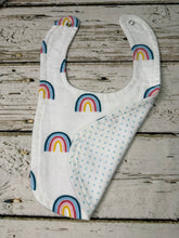 Load image into Gallery viewer, A white baby bib with a colorful rainbow pattern and a light blue dotted reverse side, laid flat on a rustic white-washed wooden surface.