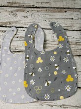 Load image into Gallery viewer, Two fabric baby bibs laid flat on a rustic whitewashed wood surface. The left bib is light gray with a white daisy print, and the right bib is medium gray with a print of yellow honeycombs, white daisies, and bumblebees.