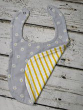 Load image into Gallery viewer, A baby bib laid flat on a rustic white-painted wooden surface. The bib has a light gray front with small white daisy flowers and a folded back panel showing yellow and white vertical stripes.