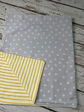 Load image into Gallery viewer, A folded fabric blanket shown on a rustic whitewashed wood surface. The main visible side is light gray with a small repeating white daisy pattern, and the folded corner reveals a yellow-and-white striped reverse side.