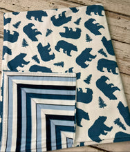 Load image into Gallery viewer, A folded baby blanket with a blue bear and tree print on an off-white background, shown next to the reverse side which features a blue and white striped geometric pattern, laid flat on a rustic wooden surface.