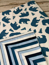 Load image into Gallery viewer, A folded blanket with a cream background and dark blue bear and tree print, shown alongside another matching piece with blue, navy, white, and light blue stripes, laid on a rustic wooden surface.