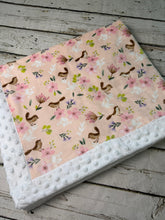 Load image into Gallery viewer, A folded baby blanket with a light pink floral and bird pattern, finished with a white minky dot border, displayed on a rustic gray wooden surface.
