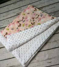 Load image into Gallery viewer, A folded baby blanket with a white minky dot textured side and a light pink floral print side, displayed on a rustic white-washed wooden surface.
