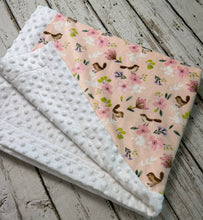 Load image into Gallery viewer, A folded baby blanket with a white minky dot side and a peach floral side with pink flowers, green leaves, and brown birds, displayed on a rustic white-washed wooden floor.
