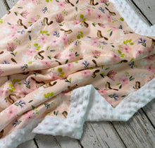 Load image into Gallery viewer, A baby blanket with a pink floral and squirrel print, featuring a white minky dot reverse side, draped on a rustic white-washed wooden background.
