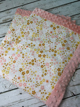 Load image into Gallery viewer, A folded baby blanket featuring a white fabric body with a pink and gold floral pattern, finished with a dusty pink minky dot border, displayed on a rustic white-washed wooden surface.
