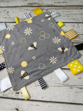 Load image into Gallery viewer, A gray baby lovey blanket with a bee and daisy pattern, featuring yellow, white, and black sensory tabs on a rustic white wood background.
