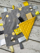 Load image into Gallery viewer, A baby sensory blanket made from fabric with a yellow minky on back, bumblebee and daisy print, and ribbon tabs in yellow, white, black, and gray, photographed on a rustic whitewashed wood surface.

