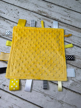Load image into Gallery viewer, A yellow minky dot sensory lovey or baby blanket with multiple fabric tabs in yellow, white, black, and striped patterns, laid flat on a rustic white wood plank surface.
