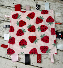Load image into Gallery viewer, A square baby sensory lovey or pacifier clip made of light pink fleece with a red strawberry pattern and colorful ribbon tabs, laid flat on a rustic white-washed wooden surface.
