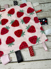 Load image into Gallery viewer, A soft baby sensory lovey featuring a light pink fleece body with a red strawberry pattern and multiple colorful ribbon tabs.
